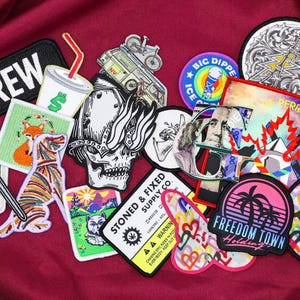 Puede incluir: Un conjunto de parches bordados con varios diseños, incluyendo una calavera, un zorro, una espada y el texto "CREW". Otros parches presentan una carita sonriente, una palmera y el texto "FREEDOM TOWN". Los parches están sobre un fondo burdeos.