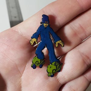 May include: A small enamel pin featuring a cartoon character wearing a dark blue beanie, a matching blue jumpsuit, and bright lime green roller skates. The character has tan skin and is depicted in a dynamic pose. A metal ruler is partially visible.