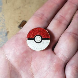May include: A round, red and white enamel pin with a glittery red top half and a white bottom half. The pin features a black horizontal line separating the two colors and a black circle in the center.