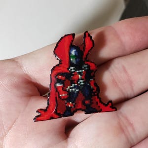 May include: A small enamel pin featuring a pixel art character design. The figure wears a dark blue suit with a prominent, flowing bright red cape that extends behind the shoulders. The character has a masked face with a green and white visor detail.