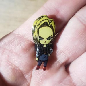 May include: A colorful pin featuring a cartoon character with blonde hair and green eyes. The character is wearing a black jacket, blue pants, and red boots. The pin is likely made of metal and has a glossy finish.