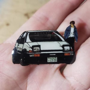 Puede incluir: Un pin de coche blanco y negro con una matrícula japonesa que dice "17-71". El pin presenta una caricatura de una persona de pie junto al coche. La palabra "TRUENO" está impresa en el coche.