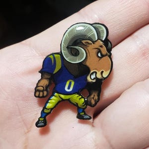 May include: A colorful pin featuring a cartoon ram in a blue and yellow football uniform with the number 0. The ram has large, curved horns and a determined expression. The pin is likely a collectible or a fan item.