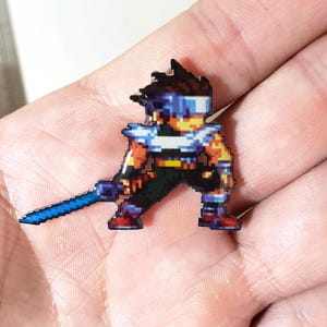May include: A pixel art enamel pin featuring a character with spiky brown hair, a blue visor, and a white scarf. The character wears dark clothing and holds a long, glowing blue sword. The design uses a blocky, retro video game aesthetic.