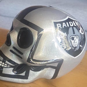 Op de afbeelding: Een zilverkleurig geschilderd schedelbeeld met zwarte accenten en een Raiders-teamlogo-sticker aan de zijkant. De schedel heeft holle oogkassen, een neusholte en een geschilderde grijns, waarbij het Raiders-schildembleem duidelijk zichtbaar is.