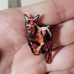 May include: A small enamel pin featuring a graphic illustration of a snarling, undead dog with exposed muscle and bone textures in shades of red, orange, and black. The dog is depicted in a running pose with pointed ears and glowing white eyes.