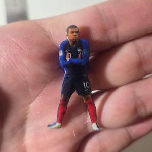 Puede incluir: Un pequeño pin plano troquelado que muestra a un futbolista con una camiseta azul oscuro con el número 10, pantalones cortos azul oscuro y calcetines rojos. El jugador aparece con los brazos cruzados y viste la equipación de la selección francesa.