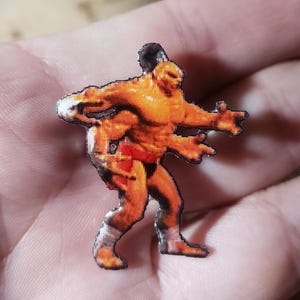 May include: A small, pixelated figurine of a muscular, orange-skinned humanoid with four arms and red shorts. The figure has a black outline and is standing with its arms outstretched.