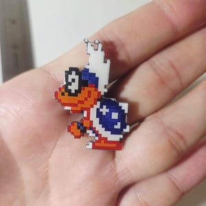 Puede incluir: Un pin de esmalte estilo pixel art que representa al personaje Lakitu de la serie Super Mario. El personaje tiene una cara naranja, gafas de sol negras, un tocado blanco en forma de nube y un caparazón azul con detalles blancos en estilo 8-bit.