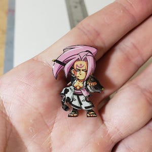 May include: A small enamel pin featuring a stylised anime character with spiky pink hair, a purple forehead mark, and a white and black outfit. The character has a determined expression and is posed in a fighting stance with one arm extended forward.