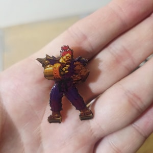 May include: A small enamel pin featuring a pixelated character with spiky red hair, wearing a dark purple martial arts uniform with a black belt, and large gold wrist cuffs, depicted in a dynamic fighting stance with one arm extended forward.