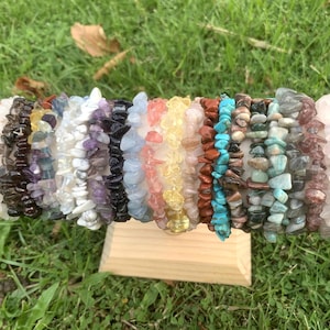 Pode incluir: Uma exibição de pulseiras de lascas de pedras preciosas coloridas. As pulseiras são feitas de várias pedras, incluindo laranja, preto, branco, roxo, azul, amarelo e verde. Elas são exibidas em um pequeno suporte de madeira sobre grama verde.