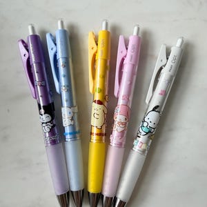 May include: Five Pilot Opt. retractable ballpoint pens featuring Sanrio character graphics. The pens are purple, light blue, yellow, light pink, and white, each with a matching clip, a clear barrel, and a silver-toned metal tip and click mechanism.