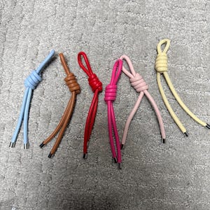 May include: A collection of six colorful leather cord loops. The loops are in various colors, including light blue, brown, red, pink, light pink, and yellow. Each loop has a knot at the top and metal tips on the ends of the cords.