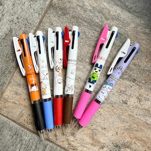 May include: A set of six multi-color ballpoint pens featuring various character designs. The pens include orange, white, red, brown, pink, and light purple barrels with matching clips and black ink tips, showcasing Peanuts and Chiikawa graphics.