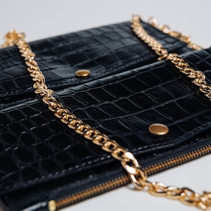 Könnte beinhalten: Schwarze Geldbörse mit goldener Kette. Die Geldbörse hat ein strukturiertes, krokodilartiges Muster und goldfarbene Hardware, einschließlich eines Reißverschlusses und Druckknöpfen. Die Kette ist dick und verleiht dem Accessoire Eleganz.