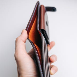 May include: Open brown leather wallet with orange interior and blue stitching. The wallet has multiple card slots and a bill compartment. The leather appears to be high-quality and well-crafted, with a classic design.