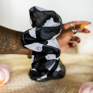 Op de afbeelding: Zwart-witte scrunchie met een herhalend spookpatroon. De scrunchie is gemaakt van een transparante stof en is gedraaid. Op de achtergrond een roze pompoen en een hand met ringen.