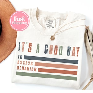 Pode incluir: Camiseta creme com o texto "IT'S A GOOD DAY TO ASSESS BEHAVIOR" em uma fonte desgastada. O design inclui listras horizontais em tons de azul, laranja e verde. Um colar de ouro com um pingente repousa sobre a camiseta.