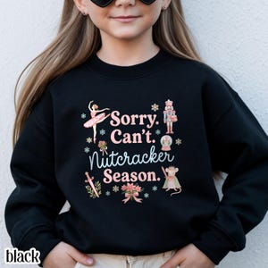 May include: A black long-sleeve sweatshirt featuring a graphic print with the text "Sorry. Can't. Nutcracker Season." The design includes illustrations of a ballerina, a nutcracker, a snow globe, a sword, a mouse, and floral accents in pink and blue.