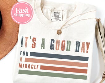 May include: Cream-colored t-shirt with the phrase "IT'S A GOOD DAY FOR A MIRACLE" in distressed lettering. The design includes horizontal stripes in muted colors. A gold necklace with a pendant rests on the shirt.
