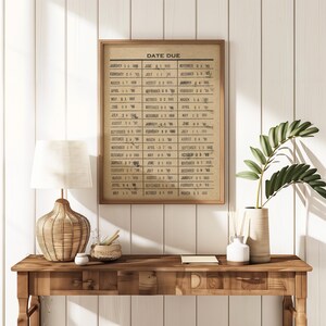 May include: A rectangular framed wall art piece featuring a tan background with the text DATE DUE at the top, followed by a grid of three columns listing various months, days, and years in black ink, presented in a simple wooden frame.