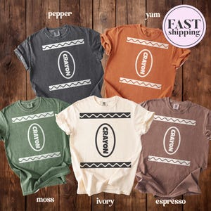 May include: Five short-sleeved crew neck t-shirts in pepper, yam, moss, ivory, and espresso colours. Each shirt features a graphic print of a crayon wrapper design with the word CRAYON inside an oval and wavy lines above and below the text.