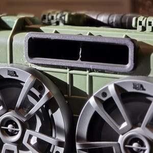 Puede incluir: Primer plano de una robusta caja de altavoz verde oliva con dos altavoces JBL plateados. Los altavoces tienen un diseño distintivo con múltiples radios. Una ventilación rectangular es visible encima de los altavoces, y la caja tiene un asa.