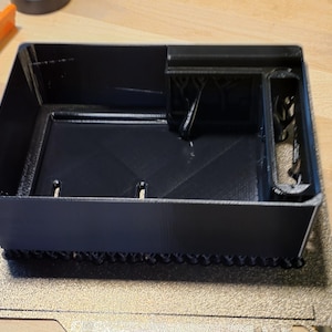 May include: A black, rectangular 3D-printed box with a divided interior. The box has a textured base and a smooth, matte finish. The design includes compartments and structural supports, suggesting a storage or organizational function.