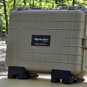 May include: A tan Apache 4800 rugged mobility case, supported by two black stands. The case features a black rectangular label and a grid pattern. The background is a blurred outdoor scene. The case has a handle on the side.