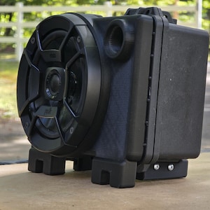 May include: A black speaker with a circular grill and a rectangular body. It features a cylindrical port and is supported by four black feet. The speaker is on a light brown surface.