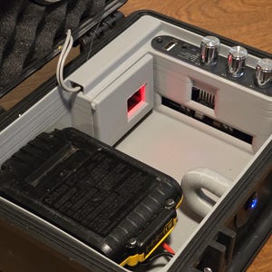 Può includere: Custodia nera aperta con componenti interni grigi. Un pacco batteria nero con accenti gialli si trova in basso a sinistra. La custodia ha un pannello di controllo con manopole argentate e una luce rossa. È visibile una porta USB.