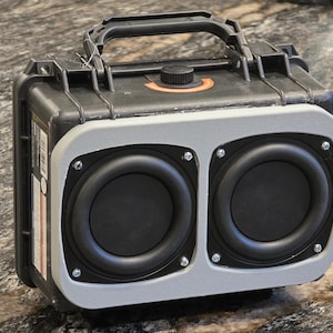 May include: A black and grey portable speaker featuring two large circular speakers. The speaker has a handle and a control knob. The speaker is encased in a robust, protective case, suggesting it's designed for outdoor use or travel.