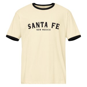Puede incluir: Camiseta color crema con ribetes negros en el cuello y las mangas. La camiseta presenta las palabras "SANTA FE" arqueadas en el pecho, con "NEW MEXICO" impreso debajo. Una prenda casual para el día a día.