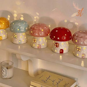 May include: A collection of ceramic mushroom-shaped lamps in various colors, including yellow, blue, red, and pink. Each lamp features a house-like base with windows and a door, and the mushroom caps are dotted with small holes that emit light. They are arranged on a white shelf.