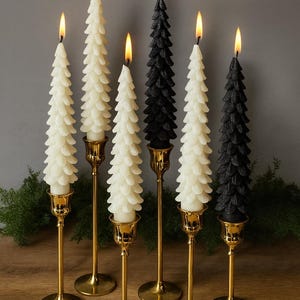 Puede incluir: Seis velas encendidas con forma de árbol en marfil y negro, colocadas en candelabros dorados. Las velas están dispuestas sobre una superficie de madera con vegetación de fondo. Las llamas son amarillas.