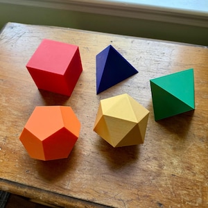 May include: A collection of five brightly colored geometric shapes: a red cube, a blue triangular pyramid, a green triangular pyramid, an orange dodecahedron, and a yellow rhombic dodecahedron. These 3D shapes are arranged on a wooden surface.