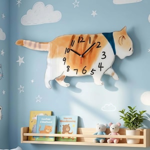 Op de afbeelding: Een grillige katvormige wandklok met een wit en oranje tabby-ontwerp. De klok heeft zwarte wijzers en zwarte cijfers. De kat draagt een blauwe kraag. Hij is gemonteerd op een lichtblauwe muur met witte wolken en sterstickers.