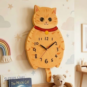 Puede incluir: Un caprichoso reloj de pared con forma de gato naranja con manecillas negras, un collar rojo y una etiqueta dorada. La esfera del reloj presenta números negros y una cara de gato sonriente.
