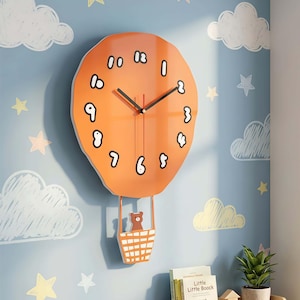 Puede incluir: Reloj de pared naranja con forma de globo aerostático con manecillas negras y números blancos. Un pequeño oso está en la cesta de abajo. El reloj está montado en una pared azul con nubes y estrellas blancas.