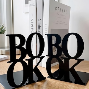 Puede incluir: Un par de sujetalibros de metal negro con las letras recortadas BOBO y OKOK. Cada sujetalibros tiene una base plana y paneles verticales con tipografía sans-serif en negrita. El diseño está construido con material negro de acabado liso.
