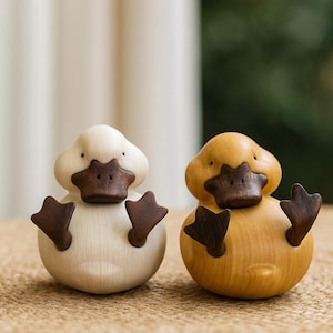 Könnte beinhalten: Zwei Holz-Entenfiguren. Eine ist cremefarben, die andere goldgelb. Beide haben dunkelbraune Schnäbel und Füße. Die Enten sitzen aufrecht, mit ausgestreckten Füßen. Der Hintergrund ist verschwommen und deutet auf eine natürliche Umgebung hin.