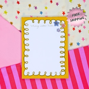 May include: A yellow notepad with a whimsical design, featuring a white interior and a decorative border. The notepad is adorned with small, colorful star-shaped embellishments and the text "Whimsy Little Notes".