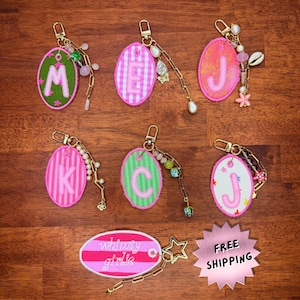 May include: A collection of personalized keychains with embroidered letters and decorative charms. Each oval-shaped tag features a different letter in pink or green, with a gold-colored chain and various beads and charms. One keychain reads "whimsy girlie".