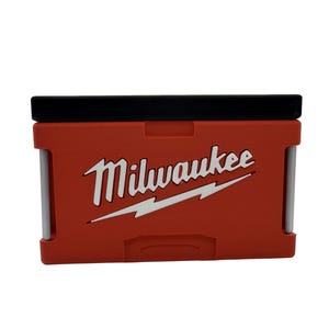 May include: A red and black Milwaukee tool box with the Milwaukee logo in white. The tool box is rectangular with a black lid and silver support bars. The Milwaukee logo includes the brand name and a white lightning bolt.