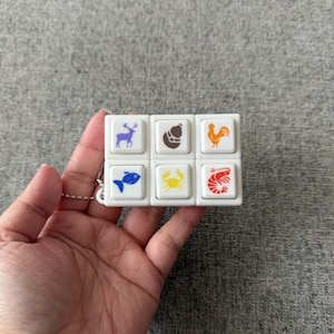 Puede incluir: Un juguete blanco en forma de cubo con seis botones cuadrados, cada uno con una ilustración de comida diferente: un ciervo, un bistec, un gallo, un pez, un cangrejo y un camarón. El juguete está unido a una pequeña cadena de metal.