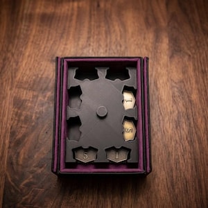 May include: A black box with a purple interior holds a black insert with cutouts for game pieces. The box contains five metallic tokens, each marked with the numbers "1" and "5". The box sits on a dark wood surface.