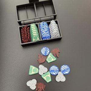 May include: A black plastic organizer box with four compartments, filled with colorful game tokens. The tokens represent fire, earth, water, and air elements. Some tokens are outside the box, scattered on a dark surface.
