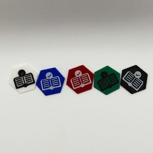 Puede incluir: Cinco fichas hexagonales de colores con un diseño de libro y marca de verificación. Las fichas son blancas, azules, rojas, verdes y negras. El libro y la marca de verificación son negros en la ficha blanca y blancos en las otras cuatro.