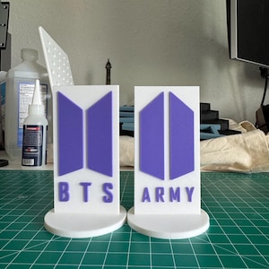 May include: Two white, 3D-printed display stands. Each stand features a purple logo and text. One reads "BTS" and the other "ARMY". The stands are on a green cutting mat.